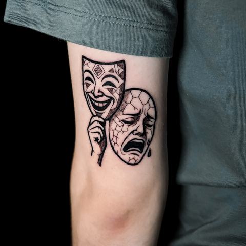 Grafisks tetovējums uz augšdelma ar teātra masku motīvu, izteiktām melnām līnijām un kontrastainu kompozīciju.