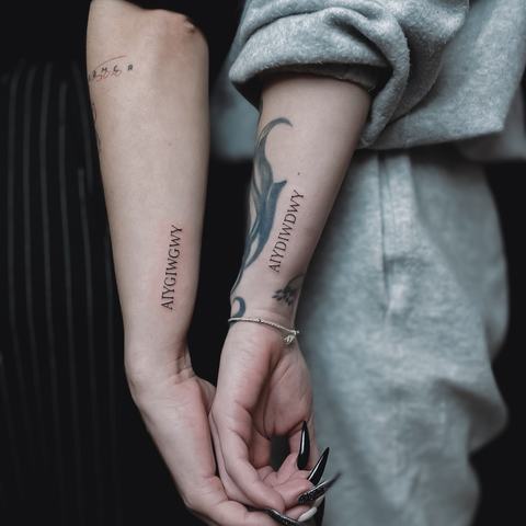 Minimalist matching forearm tattoos with delicate vertical lettering and a subtle, understated look.