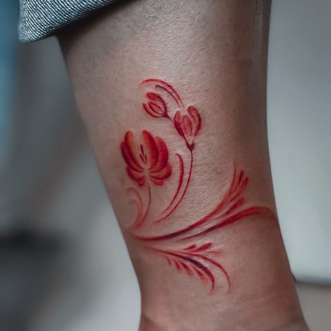 Red floral ankle tattoo with flowing stems and soft brush-like shading for a light, elegant finish.