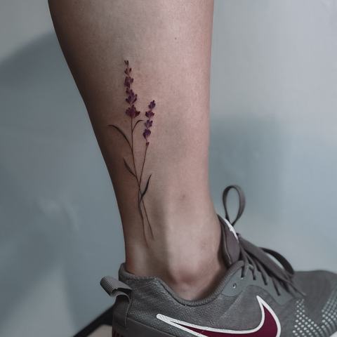 A delicate botanical tattoo along the lower leg, featuring a slender branch with fine leaves and subtle red tones.