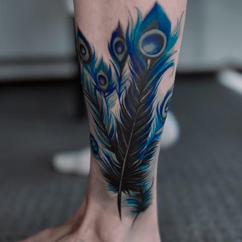 A vibrant peacock feather tattoo with deep blue and green tones, layered linework and an elegant flowing shape along the lower leg.