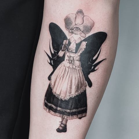 A black and grey forearm tattoo featuring a winged figure in a vintage dress, with soft shading and a slightly surreal feel.
